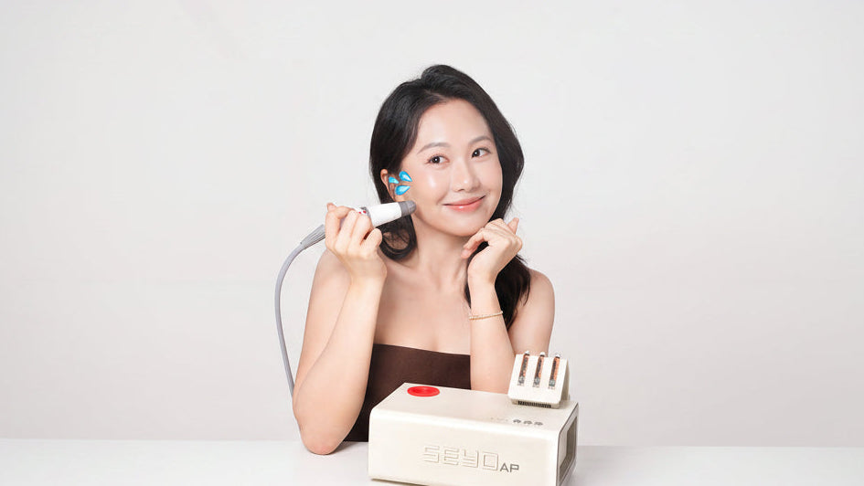 A woman holds a handheld beauty device against her ear while sitting at a white table, with a cream-colored SEYOAP beauty machine in front of her on a clean white background.